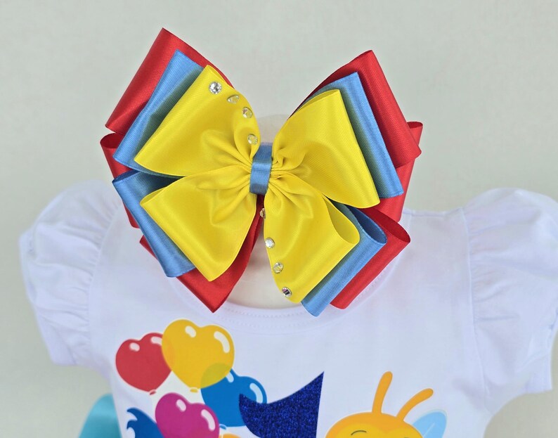 May include: A large multi-layered hair bow featuring vibrant red, blue, and yellow satin ribbons. The center is secured with a blue ribbon band, and the top yellow layer is embellished with small clear rhinestones along the side edges.
