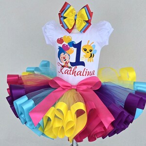 May include: A white short-sleeved shirt featuring a graphic of a clown, a bee, heart-shaped balloons, the number 1, and the name Kathalina. The shirt is paired with a multi-colored tulle tutu skirt with a large pink bow and a matching hair bow.