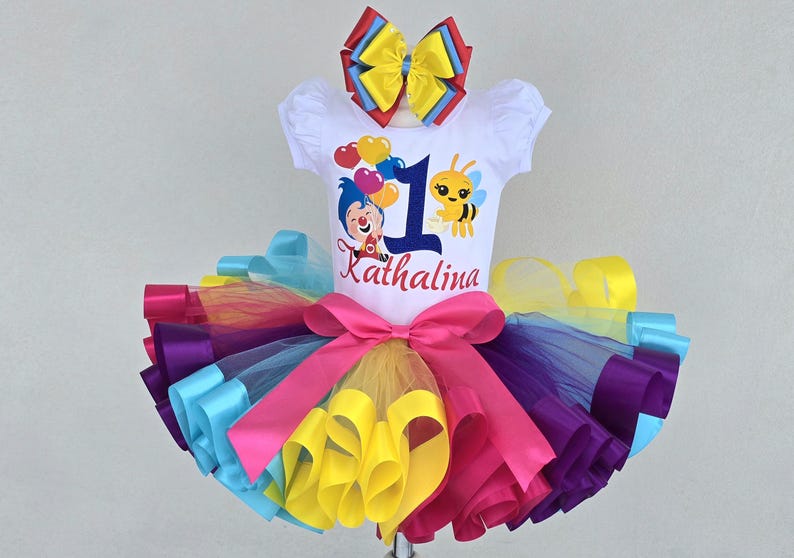 May include: A white short-sleeved shirt featuring a graphic of a clown, a bee, heart-shaped balloons, the number 1, and the name Kathalina. The shirt is paired with a multi-colored tulle tutu skirt with a large pink bow and a matching hair bow.