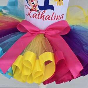 May include: A multi-colored tulle tutu skirt featuring layers of yellow, purple, and blue fabric with a bright pink satin ribbon bow at the waist. The white bodice above the skirt displays the name Kathalina in red script and a blue number one.