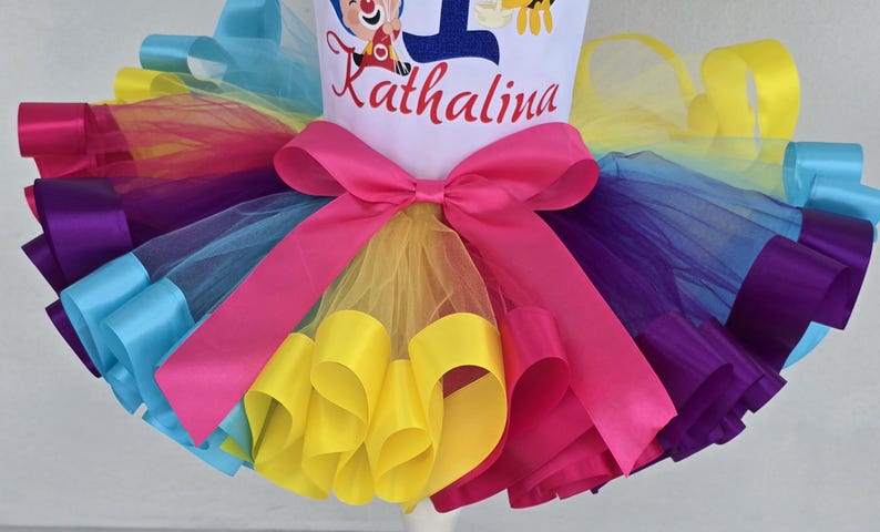 May include: A multi-colored tulle tutu skirt featuring layers of yellow, purple, and blue fabric with a bright pink satin ribbon bow at the waist. The white bodice above the skirt displays the name Kathalina in red script and a blue number one.