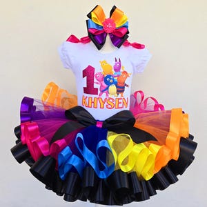 May include: A colorful birthday outfit featuring a white t-shirt with a graphic of cartoon characters and the number 1. The tutu skirt is layered with black, purple, pink, orange, yellow, and blue ribbons. A matching bow completes the ensemble.