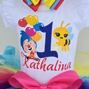 May include: A white short-sleeved t-shirt featuring a graphic of a cartoon character holding heart-shaped balloons, a yellow bee, a large dark blue glittery number one, and the name Kathalina in red script, paired with a rainbow-colored tutu.