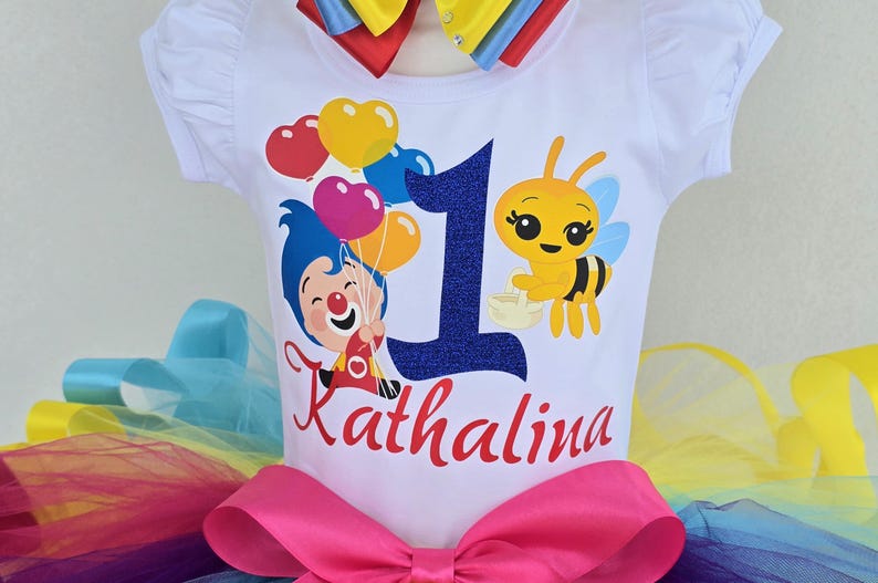 May include: A white short-sleeved t-shirt featuring a graphic of a cartoon character holding heart-shaped balloons, a yellow bee, a large dark blue glittery number one, and the name Kathalina in red script, paired with a rainbow-colored tutu.