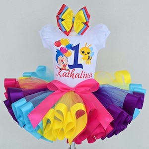 May include: A white short-sleeved bodysuit featuring a graphic of a cartoon character, a bee, and the number 1 with the name Kathalina. A multi-colored tulle tutu skirt with a large pink ribbon bow and a matching multi-colored hair bow accessory.