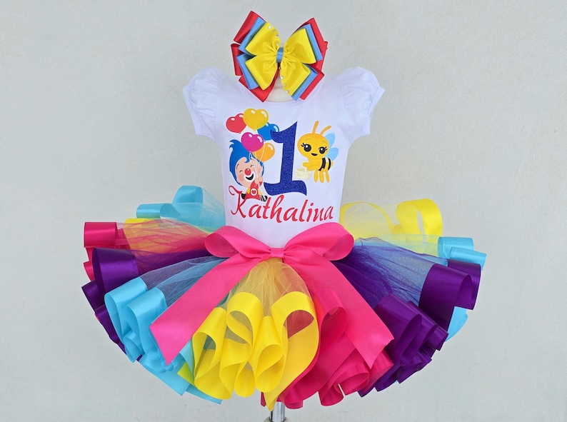 May include: A white short-sleeved bodysuit featuring a graphic of a cartoon character, a bee, and the number 1 with the name Kathalina. A multi-colored tulle tutu skirt with a large pink ribbon bow and a matching multi-colored hair bow accessory.