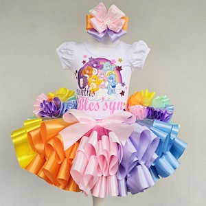 May include: A colorful rainbow-themed tutu set with a matching headband. The set includes a white t-shirt with a Care Bears graphic and the text "6 months Bles syn". The tutu features layers of yellow, orange, pink, purple, and blue ribbons.