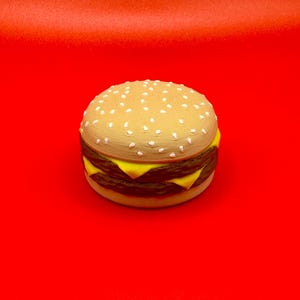 May include: A detailed image of a double cheeseburger on a red background. The burger features a sesame seed bun, two beef patties, and two slices of yellow cheese. The burger is a light tan color with white sesame seeds.