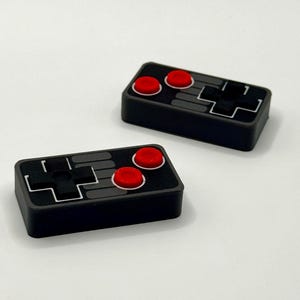 May include: Two rectangular game controller replicas in black, gray, and red. Each controller features a black directional pad, two circular red buttons with white borders, and gray rectangular accents on a textured black surface.