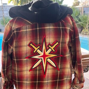 Puede incluir: Camisa de franela a cuadros rojos y tostados con una sudadera con capucha negra debajo. La camisa tiene un gran parche bordado en la espalda con una estrella con dos espadas cruzadas detrás.