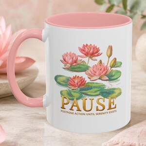 May include: A white ceramic mug with a soft pink handle and interior rim. The front features a graphic of pink lotus flowers and green lily pads. Below the flowers, the word PAUSE is printed in gold, followed by the text POSTPONE ACTION UNTIL SERENITY EXISTS.