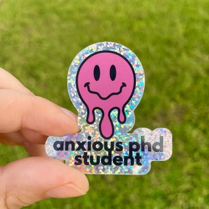 May include: A holographic sticker with a pink melting smiley face and the text "anxious PhD student".