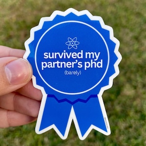 May include: Blue and white sticker with a blue ribbon. The sticker is shaped like a medal and says "survived my partner's phd (barely)" with an atom symbol in the center.
