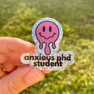 May include: A pink glitter sticker with a melting smiley face and the text "anxious phd student".