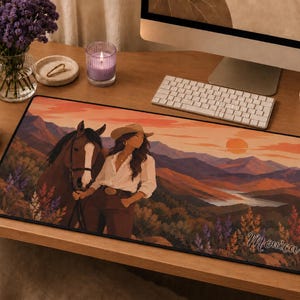 May include: A rectangular desk mat featuring a printed illustration of a person in a cowboy hat standing next to a brown horse in a mountainous landscape at sunset. The scene includes wildflowers and the name Monica written in a cursive script.