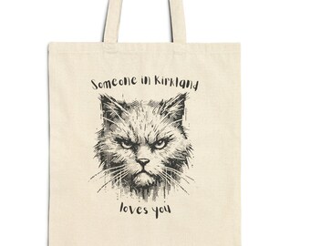 May include: Cream-colored tote bag featuring a detailed black illustration of an angry cat's face. The text "Someone in Kirkland loves you" is printed above and below the cat image. The bag has two long handles.