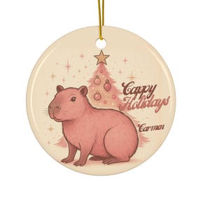 Peut inclure: Ornement de Noël rond en céramique avec un capybara rose, un sapin de Noël rose et le texte "Cappy Holidays Carmen". L'ornement a une ficelle dorée pour l'accrocher.