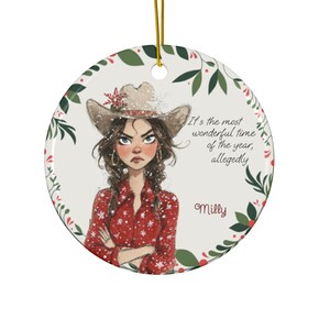 May include: A round Christmas ornament featuring a cartoon illustration of a woman wearing a cowboy hat and red shirt. The ornament has the text "It's the most wonderful time of the year, allegedly" and the name "Milly". The ornament is trimmed with green leaves and red berries.