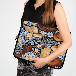 Puede incluir: Una funda negra para portátil con un diseño de vaquero, con botas, sombreros y flores azules y crema. El nombre "Rachel" está escrito en cursiva. Ideal para proteger un portátil.