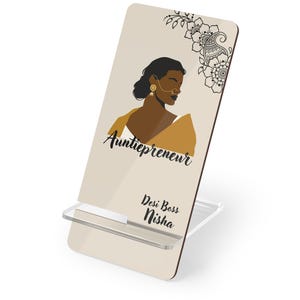 May include: A beige phone stand with a graphic of a woman wearing a nose ring and a yellow shawl. The stand has the text "Auntiepreneur" and "Desi Boss Nisha" in black script. A floral design is in the upper right corner.