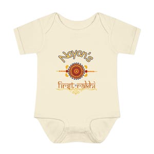 Puede incluir: Body de bebé color crema con mangas cortas. La parte delantera presenta el texto "Nayan's First Rakhi" en una fuente decorativa, junto con un diseño circular colorido. El diseño incluye elementos naranjas, amarillos y azules.