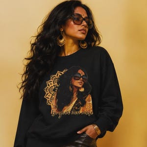 May include: A black long-sleeve sweatshirt featuring a graphic print of a person wearing sunglasses and large earrings, set against a gold mandala pattern. The text below the graphic reads "be your own muse." The sweatshirt is paired with black leather-look flared pants.