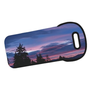 Puede incluir: Una puesta de sol púrpura y rosa sobre una silueta de un bosque. La imagen está impresa en un portador de botellas de vino de neopreno negro con un asa negra.