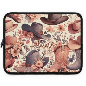 May include: A rectangular laptop sleeve featuring a repeating pattern of cowboy hats, cowboy boots, and floral arrangements in shades of peach, cream, and muted grey. The name Leanne is written in a cursive script in the center of the design.