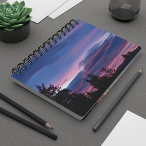 Puede incluir: Cuaderno con espiral con un diseño de puesta de sol en tonos azules, rosas y morados. La portada muestra un paisaje con árboles silueteados. Incluye lápices negros y una planta suculenta. Mide aproximadamente 21 cm de largo.