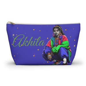 May include: A blue cosmetic bag with a colorful illustration of a woman wearing a green, blue, and red jacket and blue pants. The woman is sitting with her legs crossed and has a gold necklace and earrings. The bag has the word "Akhil" written in green cursive on the front.