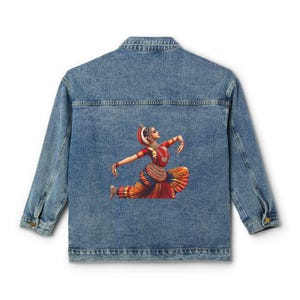 May include: Light blue denim jacket featuring a colorful illustration of an Indian dancer on the back. The dancer is depicted in a traditional pose, wearing a red and orange costume with gold jewelry. The jacket has a classic design with a button-up front and long sleeves.