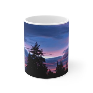 May include: A white ceramic mug featuring a printed image of a colourful sunset scene. The scene depicts a silhouette of trees against a pink, purple and blue sky.