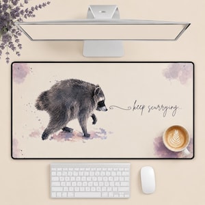 May include: A rectangular desk mat featuring a watercolor illustration of a gray and black raccoon. The text "keep scurrying..." is written in black cursive script to the right of the raccoon. The background is a light cream color with soft purple watercolor splashes.