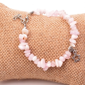 May include: A delicate bracelet featuring light pink rose quartz chips strung together. The bracelet has a silver-toned clasp and a charm with a star design. The bracelet is displayed on a tan burlap pillow.