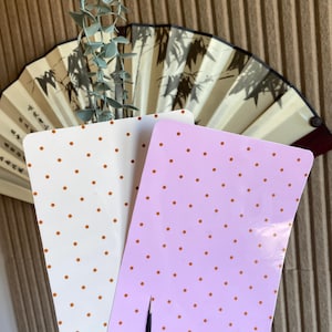 May include: Two rectangular cards with rounded corners, one white and one light pink, both featuring a repeating pattern of small orange polka dots. A thin black tool is positioned vertically against the bottom edge of the pink card.