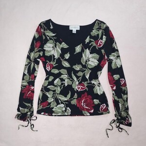 May include: Black long-sleeve top with a V-neck and a floral pattern of red roses and green leaves. The sleeves have tie details at the wrists. The brand name "ASSEX" is visible on a small tag near the neckline.