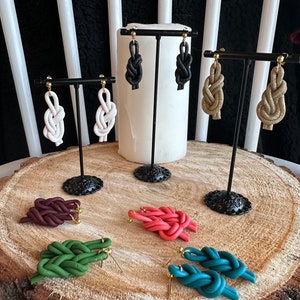 May include: Assortment of handmade knotted earrings in various colours, including white, black, beige, maroon, coral, green, and teal. The earrings are displayed on a wooden stand.