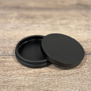 May include: A black, round container with a lid, resting on a wooden surface. The lid is slightly ajar, revealing the interior of the container. The container and lid are both matte black.