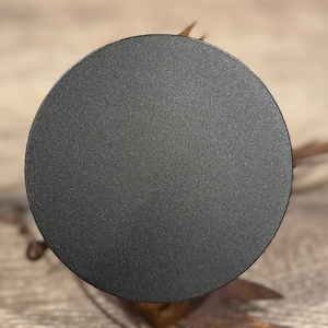 May include: A close-up shot of a matte black, circular object with a textured surface. The object is centered in the frame, with a blurred background of brown and tan elements. The object appears to be a lid or cover.