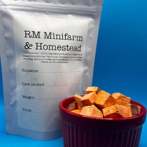 May include: A white bag with the text "RM Minifarm & Homestead" next to a small, burgundy-coloured bowl filled with orange and brown cubed food items. The bag has lines for "Date packed", "Weight", and "Price".