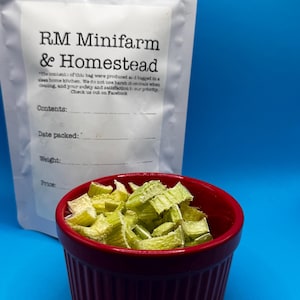May include: A red, ribbed ceramic bowl filled with chopped, light green vegetables sits in front of a white bag. The bag is labeled "RM Minifarm & Homestead" with text about the contents and Facebook. The background is a solid blue.