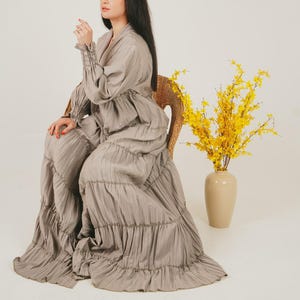 May include: A woman sits in a wicker chair wearing a long, tiered, grey dress. The dress has a ruffled neckline and long sleeves. A bouquet of yellow flowers in a beige vase is on the right side of the image.