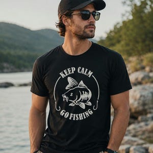 Peut inclure: Un t-shirt noir à manches courtes avec un motif blanc représentant un poisson à l'intérieur d'un cercle, accompagné du texte "KEEP CALM GO FISHING" imprimé en arc de cercle autour de l'illustration du poisson.
