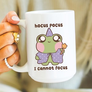 Könnte beinhalten: Weiße Keramiktasse mit einem Cartoon-Frosch-Zauberer-Design und dem Text "hocus pocus I cannot focus". Der Frosch ist grün mit rosa Wangen und einem lila Umhang. Die Tasse wird von einer Person gehalten.
