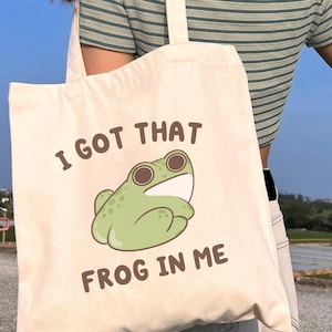 May include: Cream-colored tote bag with the text "I GOT THAT FROG IN ME" and a cartoon frog illustration. The bag has a simple design with a rectangular shape and long straps. The background is a clear blue sky.
