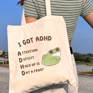 May include: A cream-colored tote bag with the text "I GOT ADHD" and a green cartoon frog illustration. The bag also has the text "Attention Deficit Hold up is Dat a Frog?" The bag is carried by a person wearing a green striped shirt.