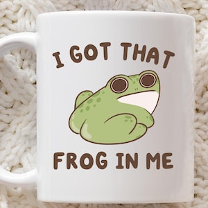 Puede incluir: Taza de cerámica blanca con una ilustración de rana verde y el texto "I GOT THAT FROG IN ME" en marrón. La taza tiene un asa curva. El fondo es de punto blanco.