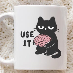 May include: White ceramic mug featuring a black cat illustration holding a pink brain. The mug has the words "USE IT" printed on the side. The mug has a white handle and is suitable for hot or cold beverages.