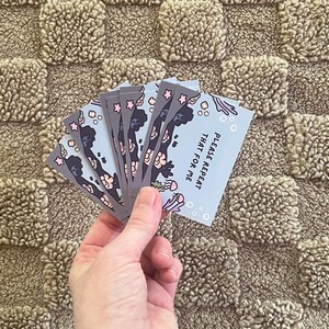 Puede incluir: Una mano sostiene una pila de tarjetas grises con un fondo azul claro. Las tarjetas presentan un dise&ntilde;o negro y rosa con el texto "PLEASE REPEAT THAT FOR ME". Las tarjetas est&aacute;n sobre una superficie marr&oacute;n texturizada.