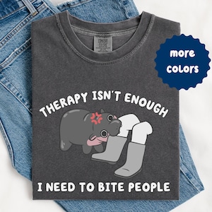 May include: Dark gray t-shirt with the text "THERAPY ISN'T ENOUGH I NEED TO BITE PEOPLE" and a cartoon hippo biting a pair of gray boots. The shirt is folded on top of blue jeans.
